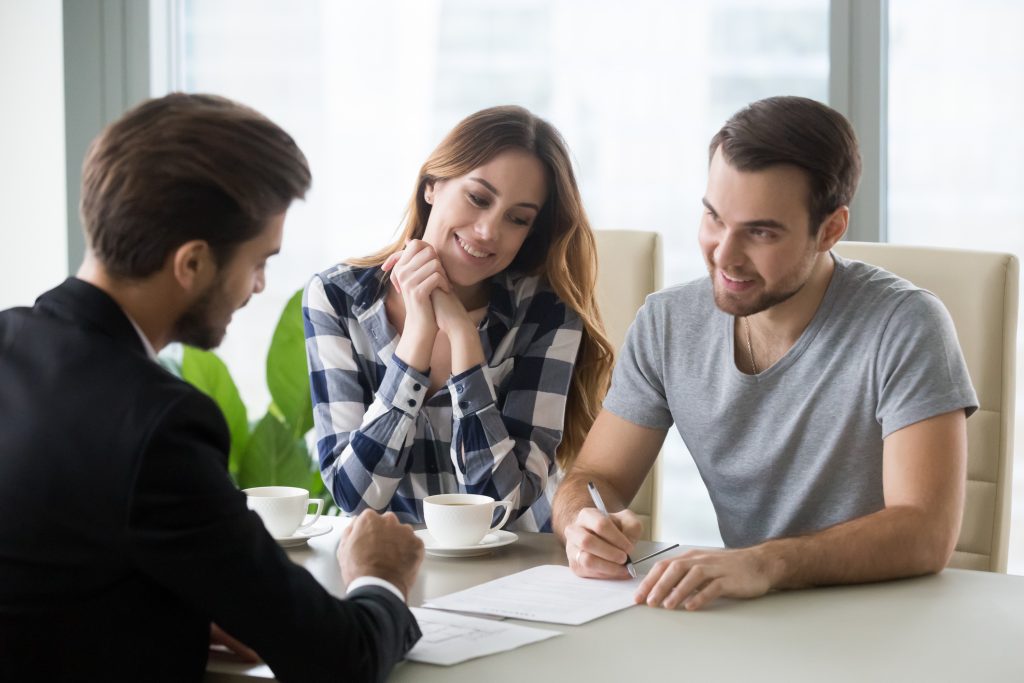 A couple is ready to sign the paperwork on their new mortgage. They're smiling as they put the pen to the paper and the mortgage advisor is offering advice.