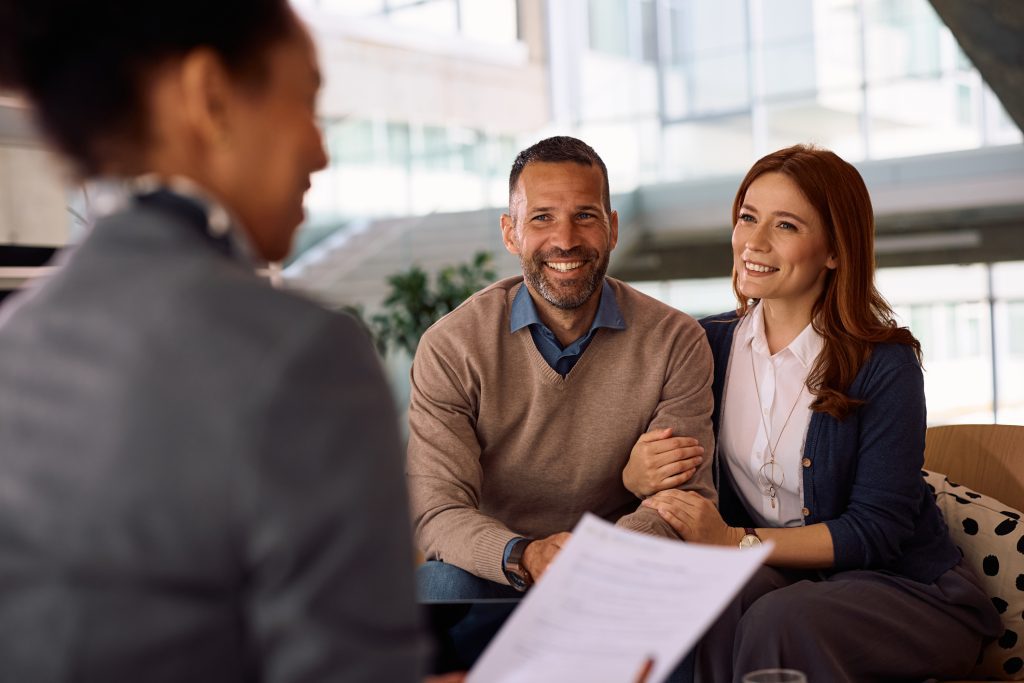 Smiling couple meeting with a professional mortgage advisor in a modern office setting, reviewing documents together. The woman is holding the man's arm affectionately while they listen attentively.